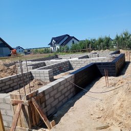 Andrii Dets - Fundamenty domu w trakcie budowy: bloczki betonowe, izolacja pionowa, szalunek. Widok na domy w tle i błękitne niebo. Prace ziemne i budowlane w toku.