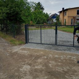 Andrii Dets - Nowa metalowa brama wjazdowa z siatki na posesję, widok z zewnątrz. Szara brama zamontowana na betonowej wylewce, teren wysypany żwirem, w tle dom z ogrodem.