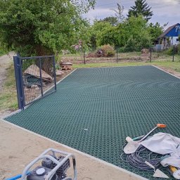 Andrii Dets - Przygotowanie terenu pod trawnik z użyciem geokraty. Na pierwszym planie ubijarka, narzędzia i piasek. Widoczne ogrodzenie i zieleń w tle.