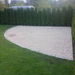 Andrii Dets - Ukończona aranżacja przestrzeni: biały żwir, obrzeża z kostki, trawnik i tuje. W tle budynek z reklamą okien i drzwi.