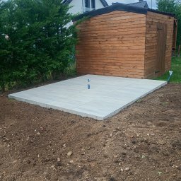 Andrii Dets - Wykonana podbudowa z jasnych płyt chodnikowych pod drewniany domek narzędziowy w ogrodzie. Widoczne niebieskie rurki. W tle dom z panelami słonecznymi.