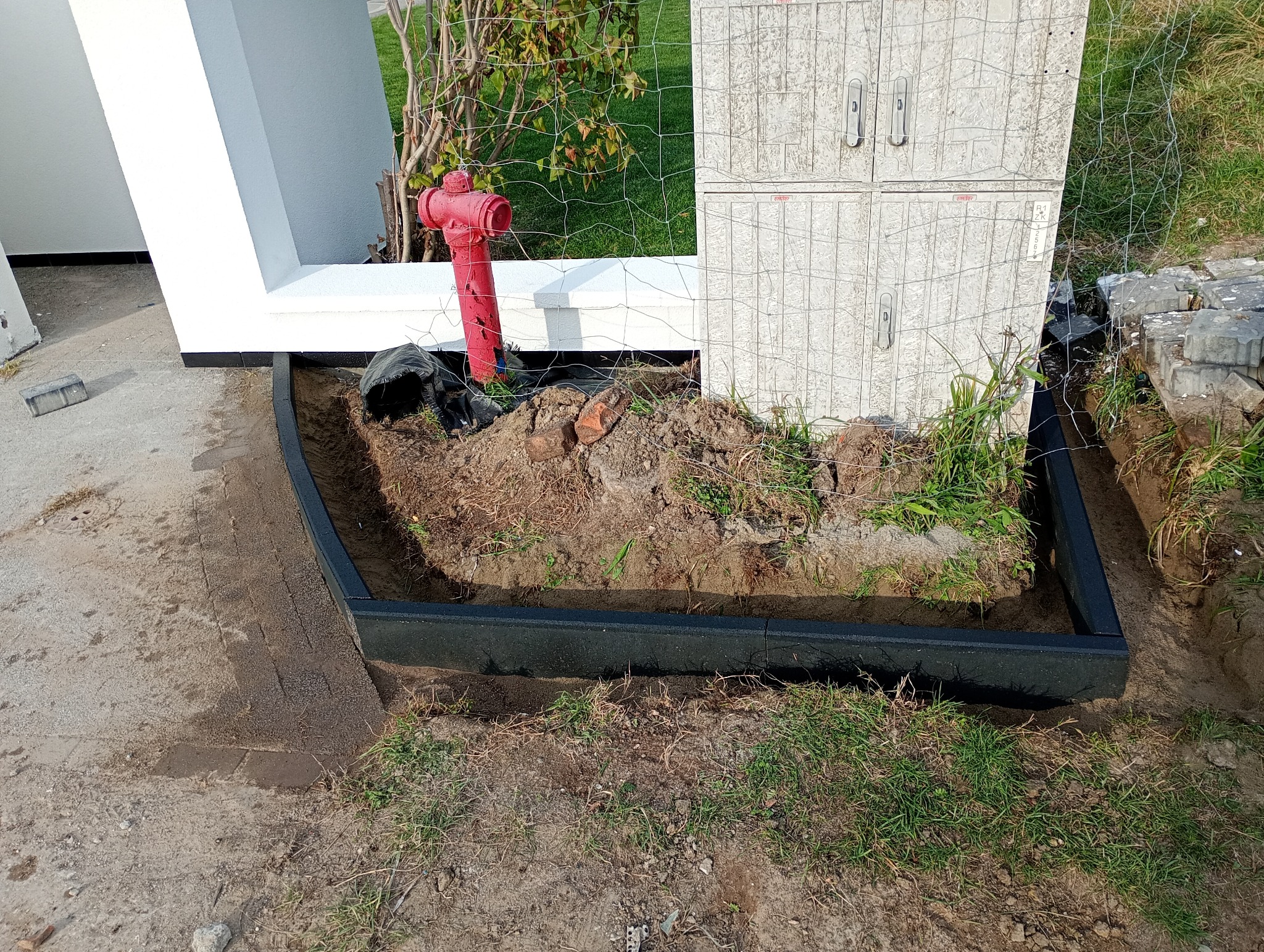 Montaż obrzeży trawnikowych z tworzywa sztucznego, widoczny fragment trawnika, hydrant i skrzynka elektryczna w tle.