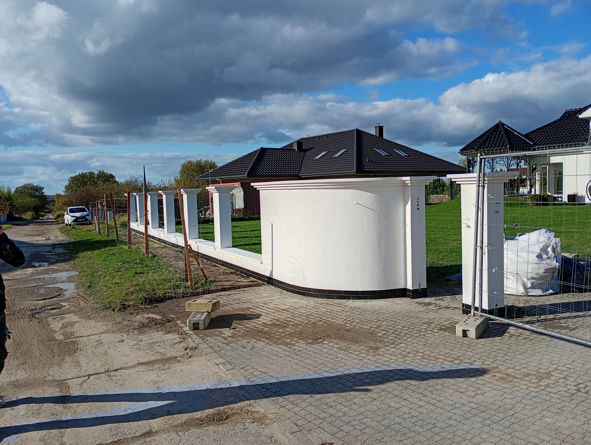 Nowo wybudowane, białe ogrodzenie z filarami i podmurówką z czarną glazurą, widoczne na tle domu z czarnym dachem i błękitnego nieba z chmurami.