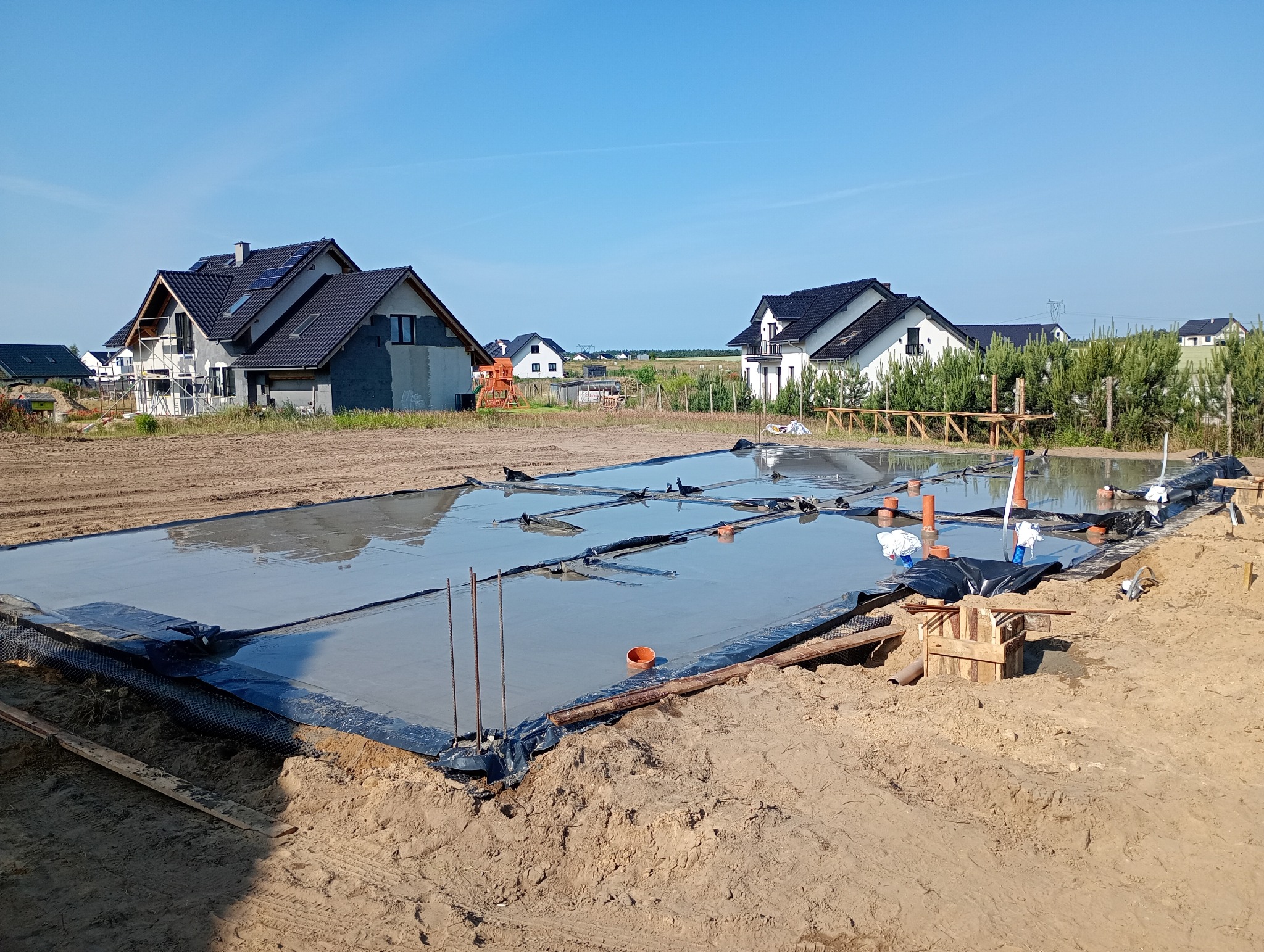 Wylany fundament na czarnej folii, otoczony piaskiem i drewnianymi elementami. W tle domy w zabudowie jednorodzinnej pod błękitnym niebem.