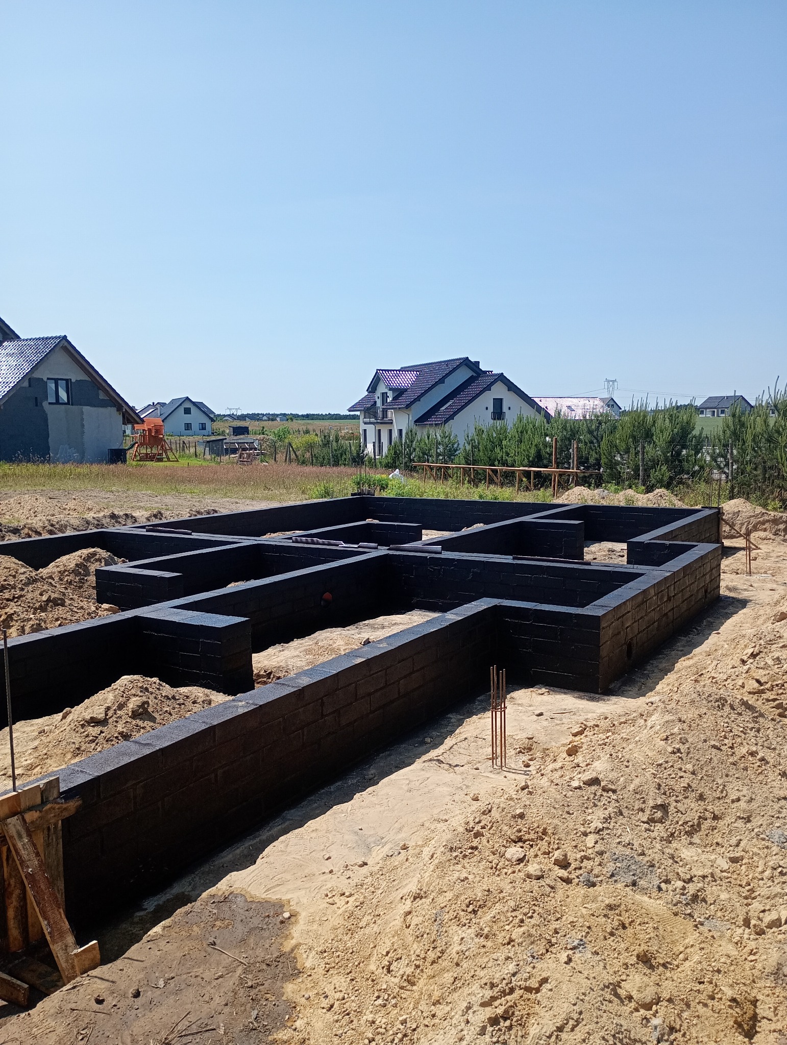 Czarna izolacja fundamentów z cegły na piasku, na tle domy jednorodzinne i błękitne niebo. Widok na solidne fundamenty domu w słoneczny dzień.