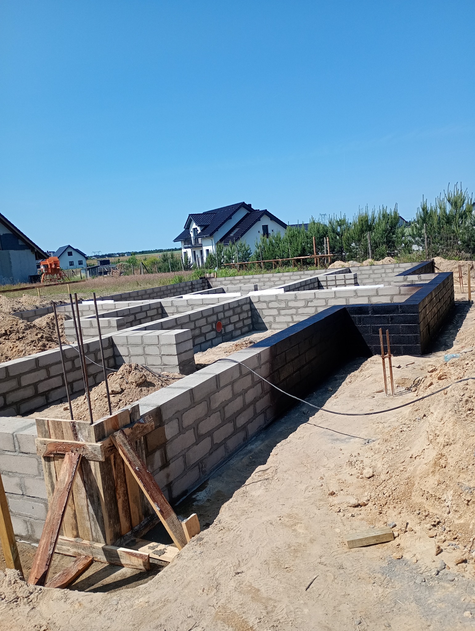 Fundamenty domu w trakcie budowy: bloczki betonowe, izolacja pionowa, szalunek. Widok na domy w tle i błękitne niebo. Prace ziemne i budowlane w toku.