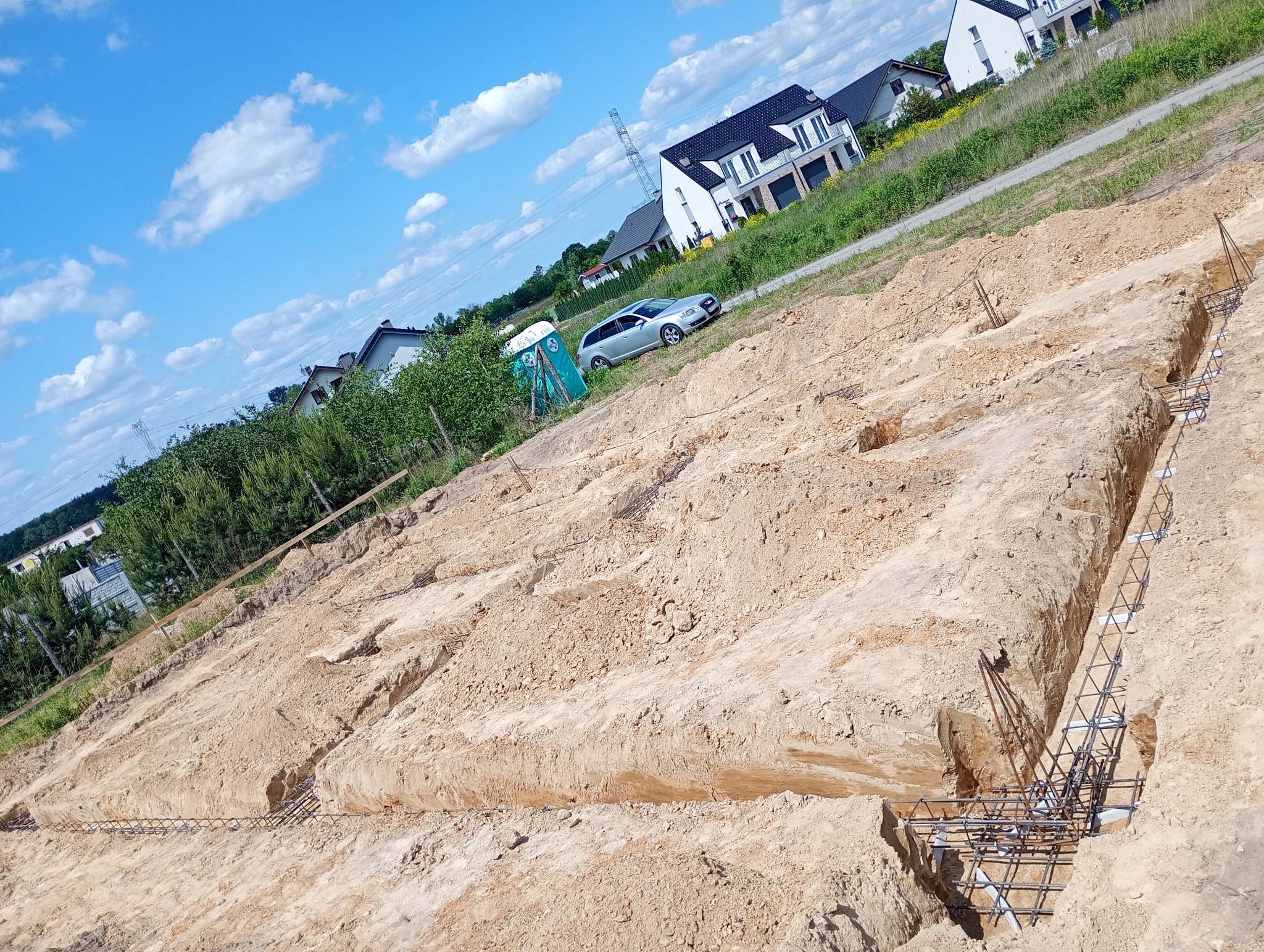 Wykop pod fundamenty z widocznym zbrojeniem, na dalszym planie domy jednorodzinne i przenośna toaleta. Ujęcie w słoneczny dzień.