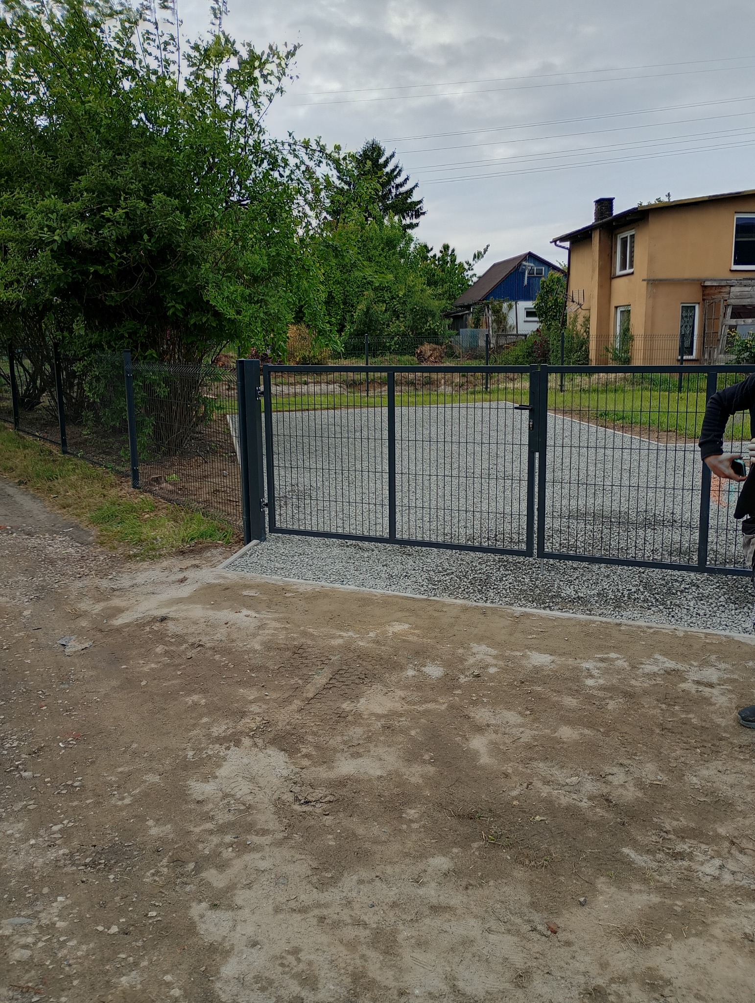 Nowa metalowa brama wjazdowa z siatki na posesję, widok z zewnątrz. Szara brama zamontowana na betonowej wylewce, teren wysypany żwirem, w tle dom z ogrodem.
