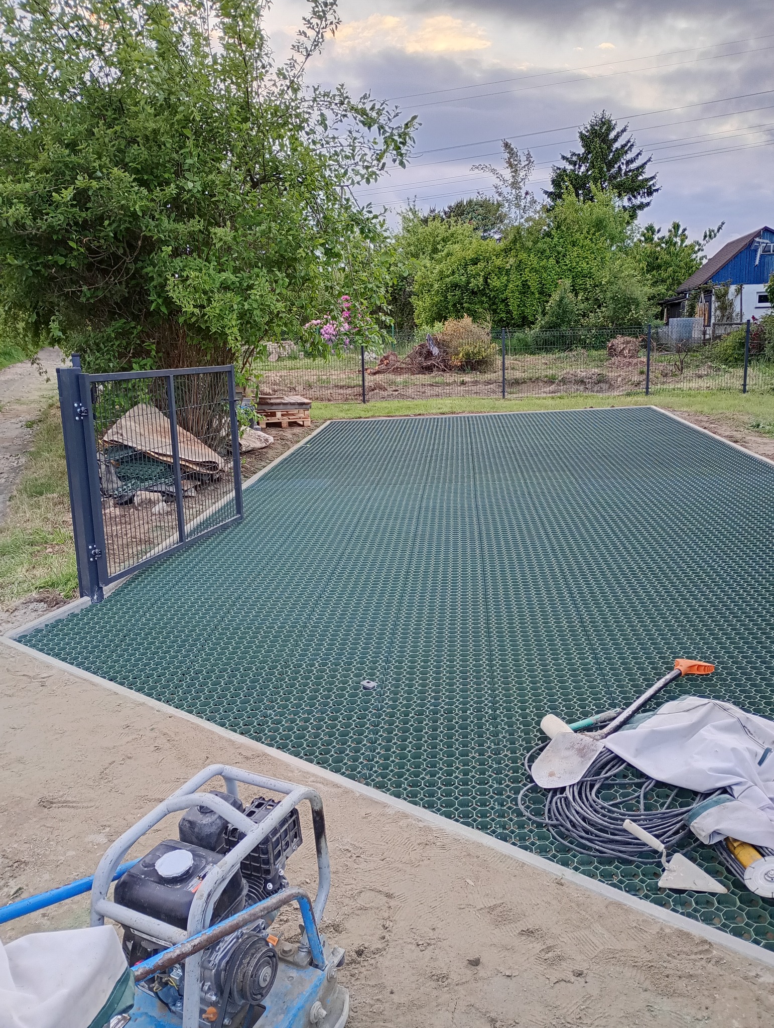 Przygotowanie terenu pod trawnik z użyciem geokraty. Na pierwszym planie ubijarka, narzędzia i piasek. Widoczne ogrodzenie i zieleń w tle.