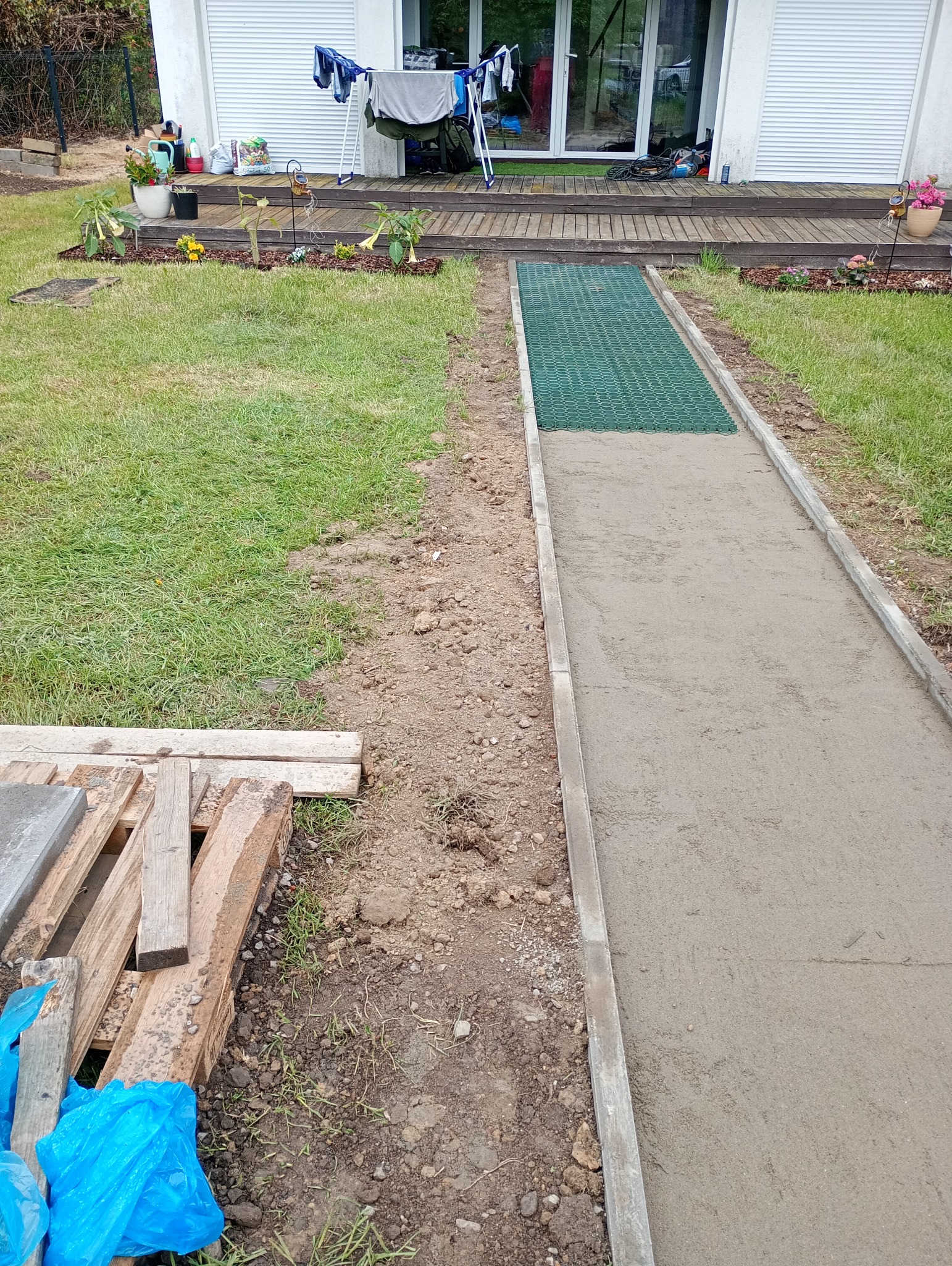 Wykonywanie ścieżki z betonową obwódką i elementami ażurowymi. Widoczne materiały budowlane i trawnik. Prace w toku, świeży beton.