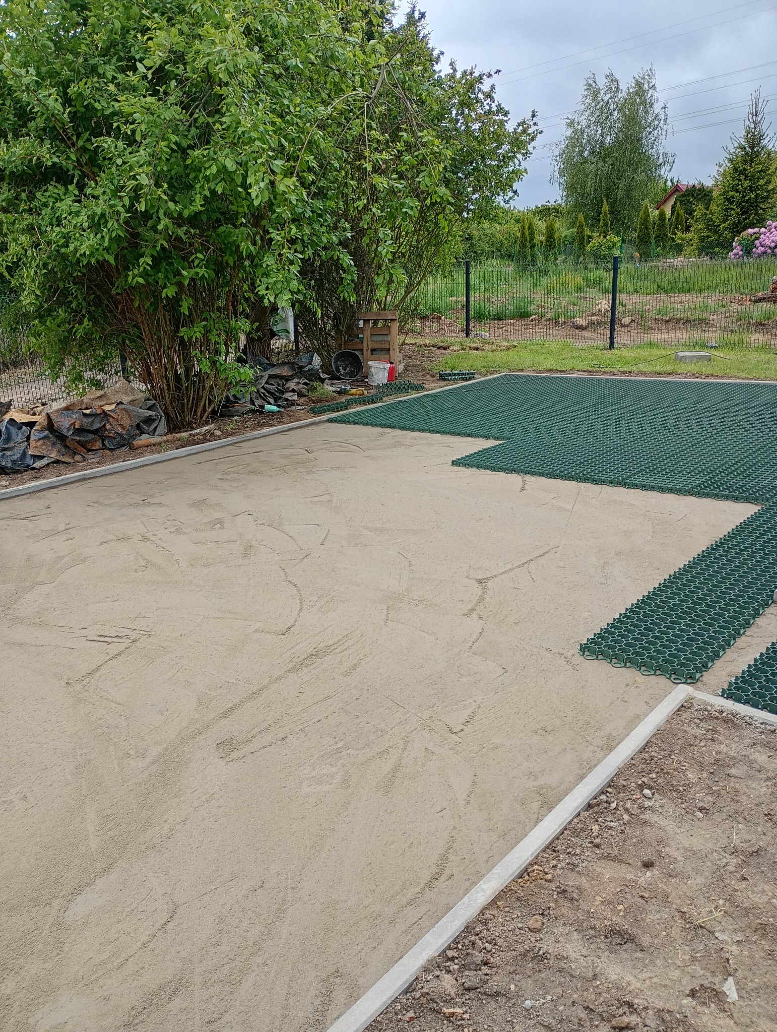 Układanie zielonych paneli trawnikowych na piaskowym podłożu, obrzeża betonowe, widok z góry, częściowo ukończony projekt, ogród w tle.