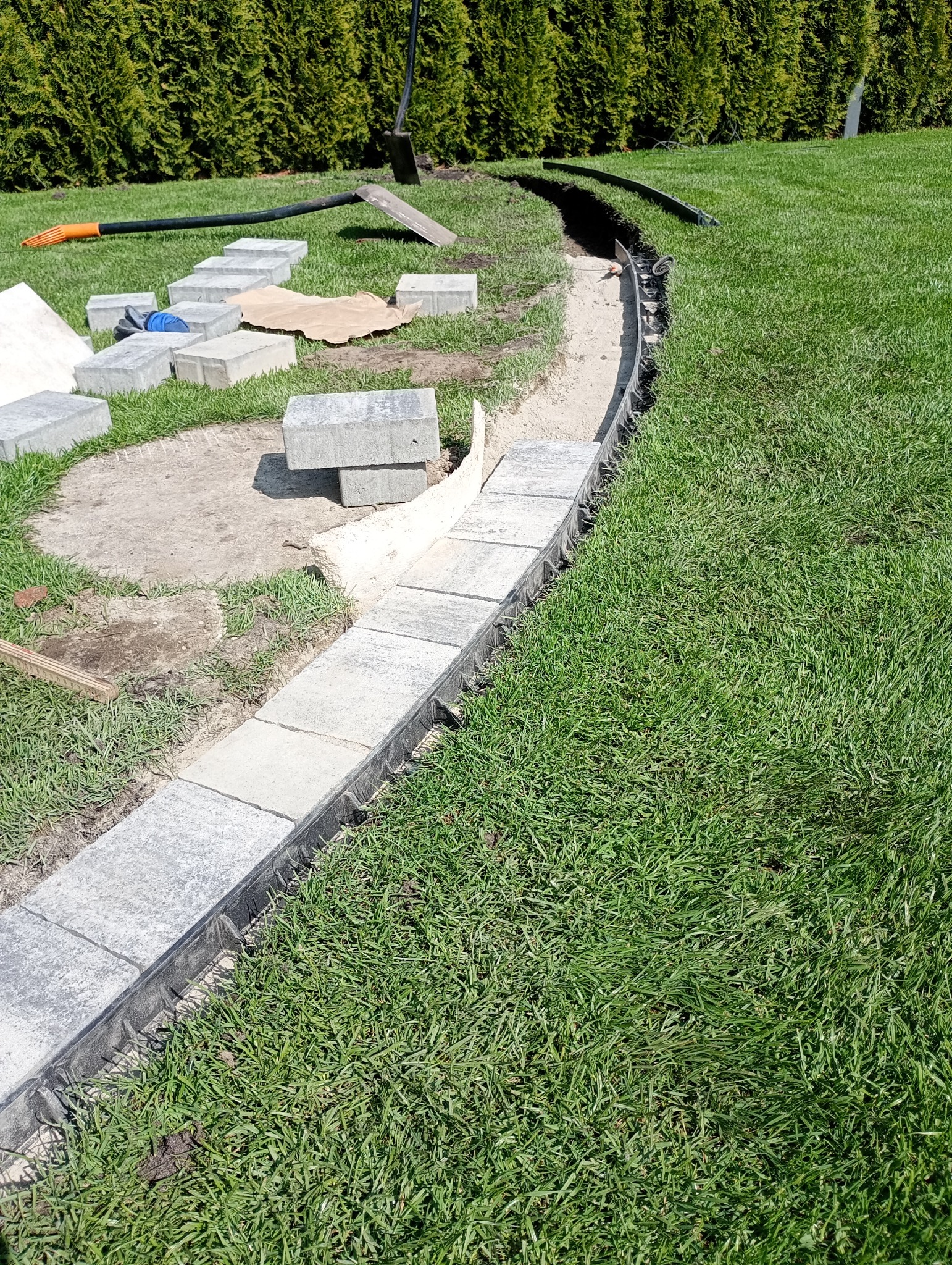 Układanie obrzeży trawnikowych z kostki betonowej. Widoczne narzędzia i materiały budowlane na trawie. Obrzeże oddziela trawnik od rabaty.