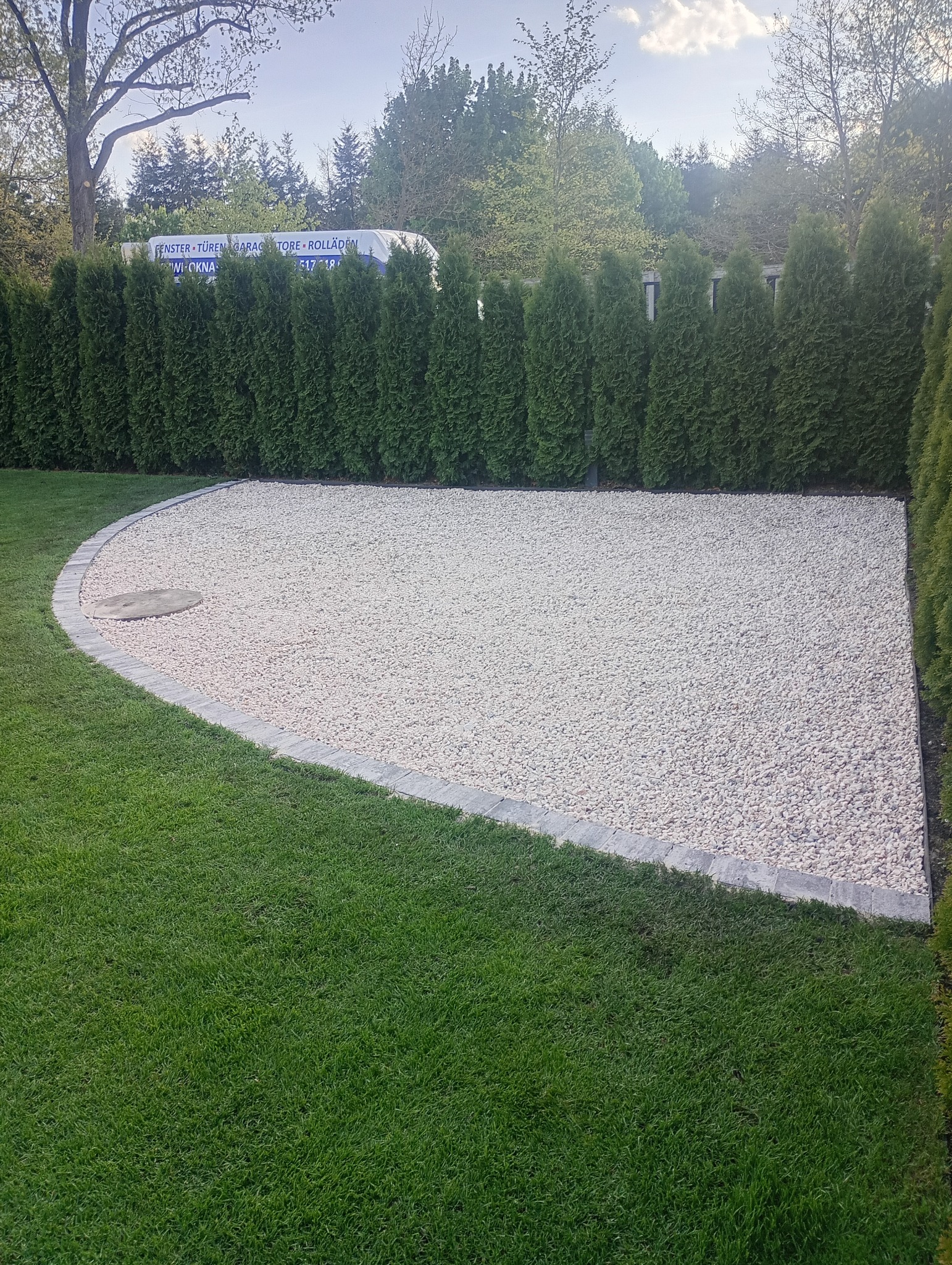 Ukończona aranżacja przestrzeni: biały żwir, obrzeża z kostki, trawnik i tuje. W tle budynek z reklamą okien i drzwi.