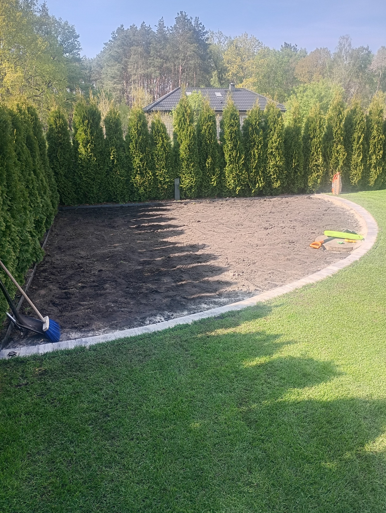 Przygotowany teren pod trawnik z obrzeżem, narzędzia ogrodnicze w rogu. W tle rząd tuj i dom. Widok z góry, słoneczny dzień.