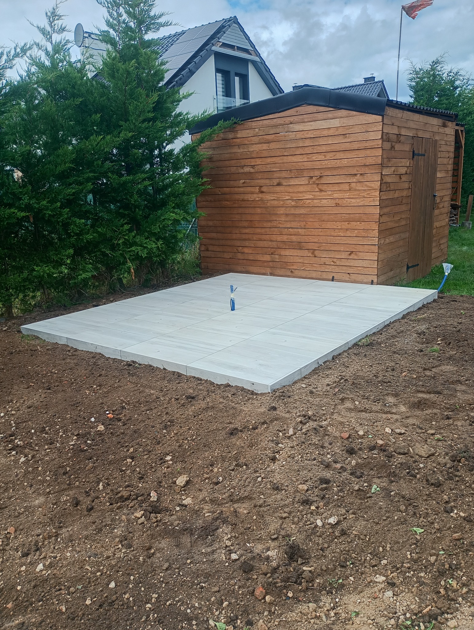 Wykonana podbudowa z jasnych płyt chodnikowych pod drewniany domek narzędziowy w ogrodzie. Widoczne niebieskie rurki. W tle dom z panelami słonecznymi.