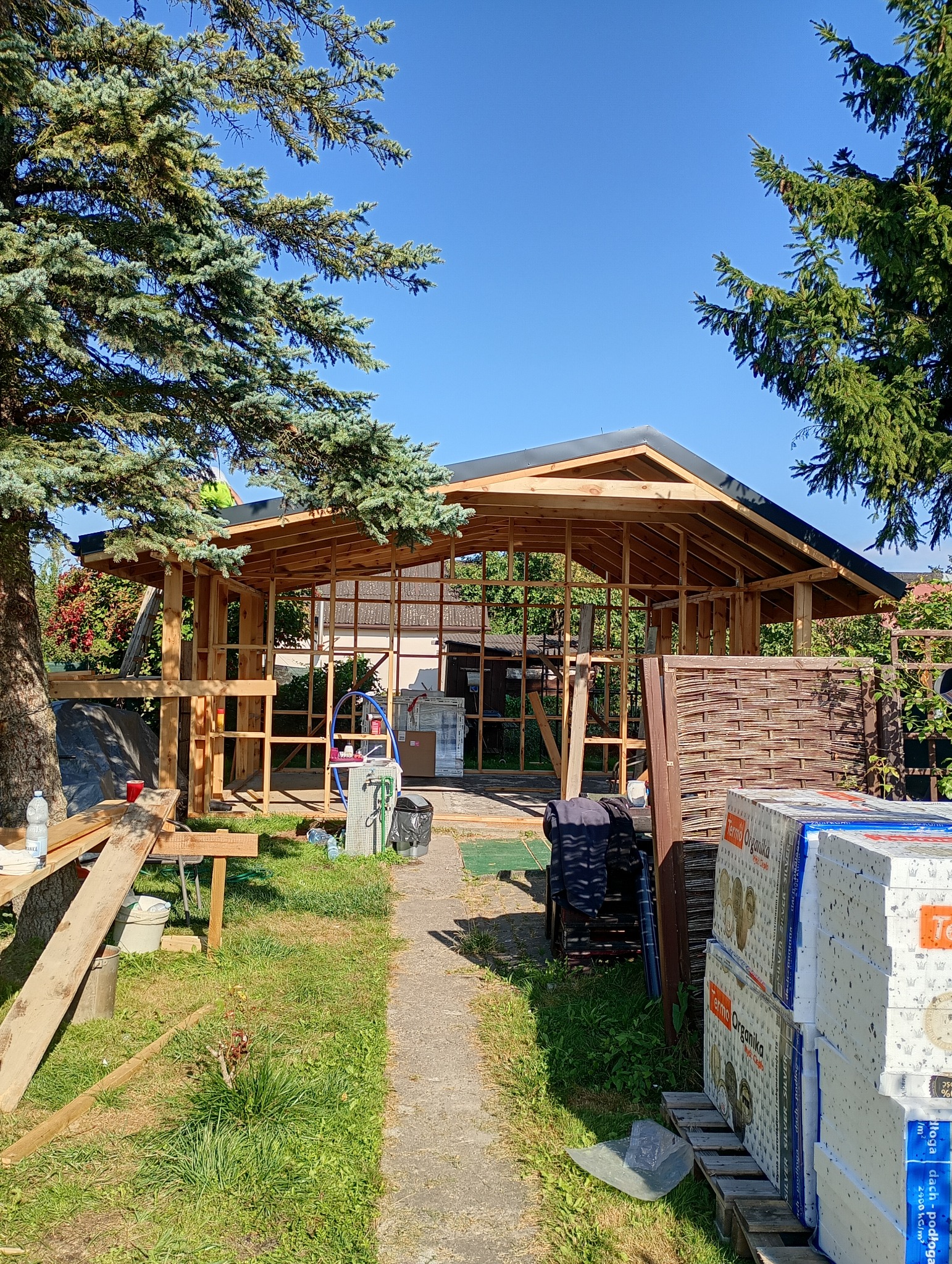 Konstrukcja szkieletowa budynku w ogrodzie, widoczna z zewnątrz. Elementy drewniane, częściowo zadaszona. W tle zieleń, drzewa i błękitne niebo.