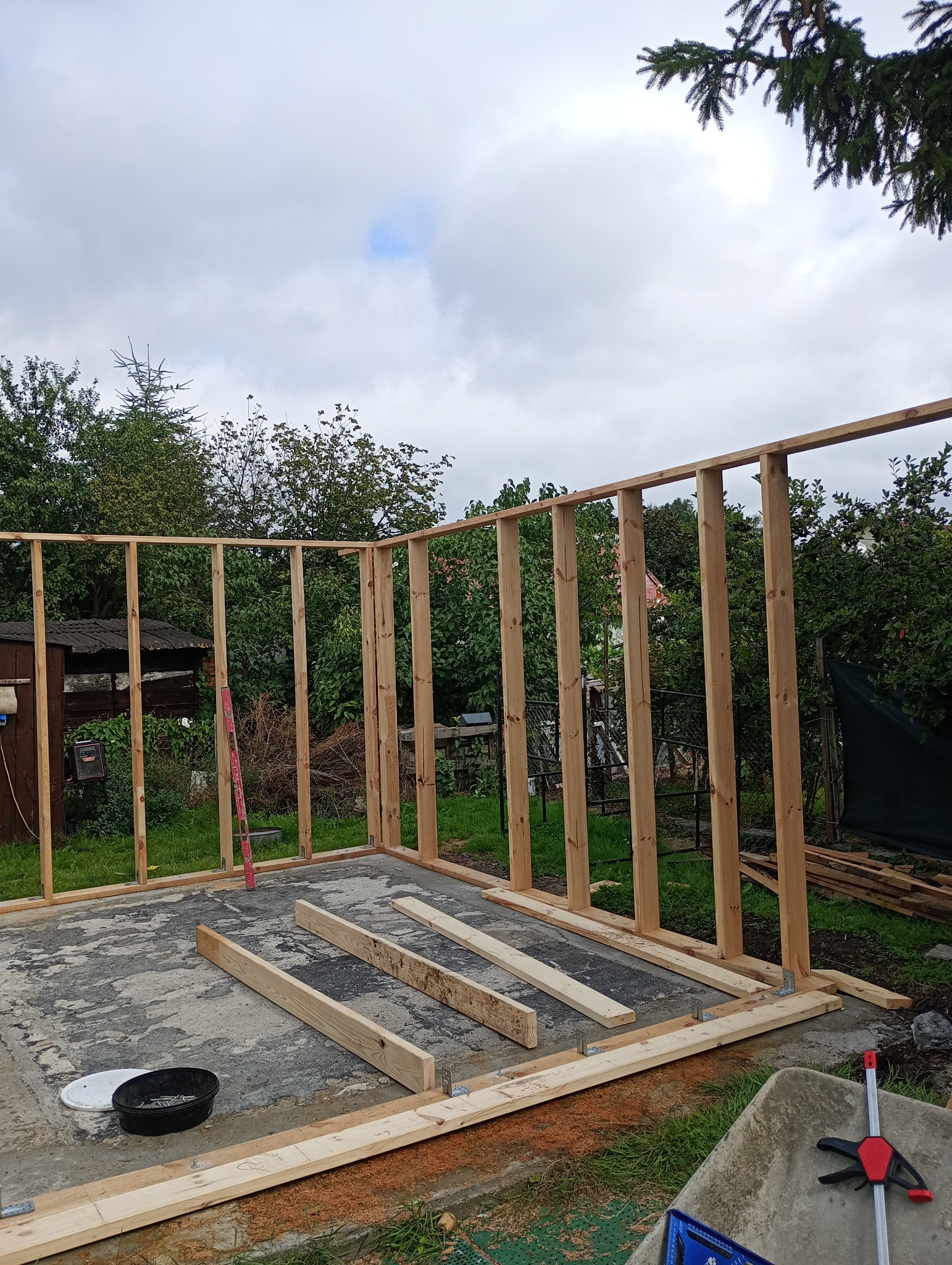Wznoszenie drewnianej konstrukcji szkieletowej na betonowej płycie, widoczne narzędzia i poziomica, w tle zieleń ogrodu i pochmurne niebo.