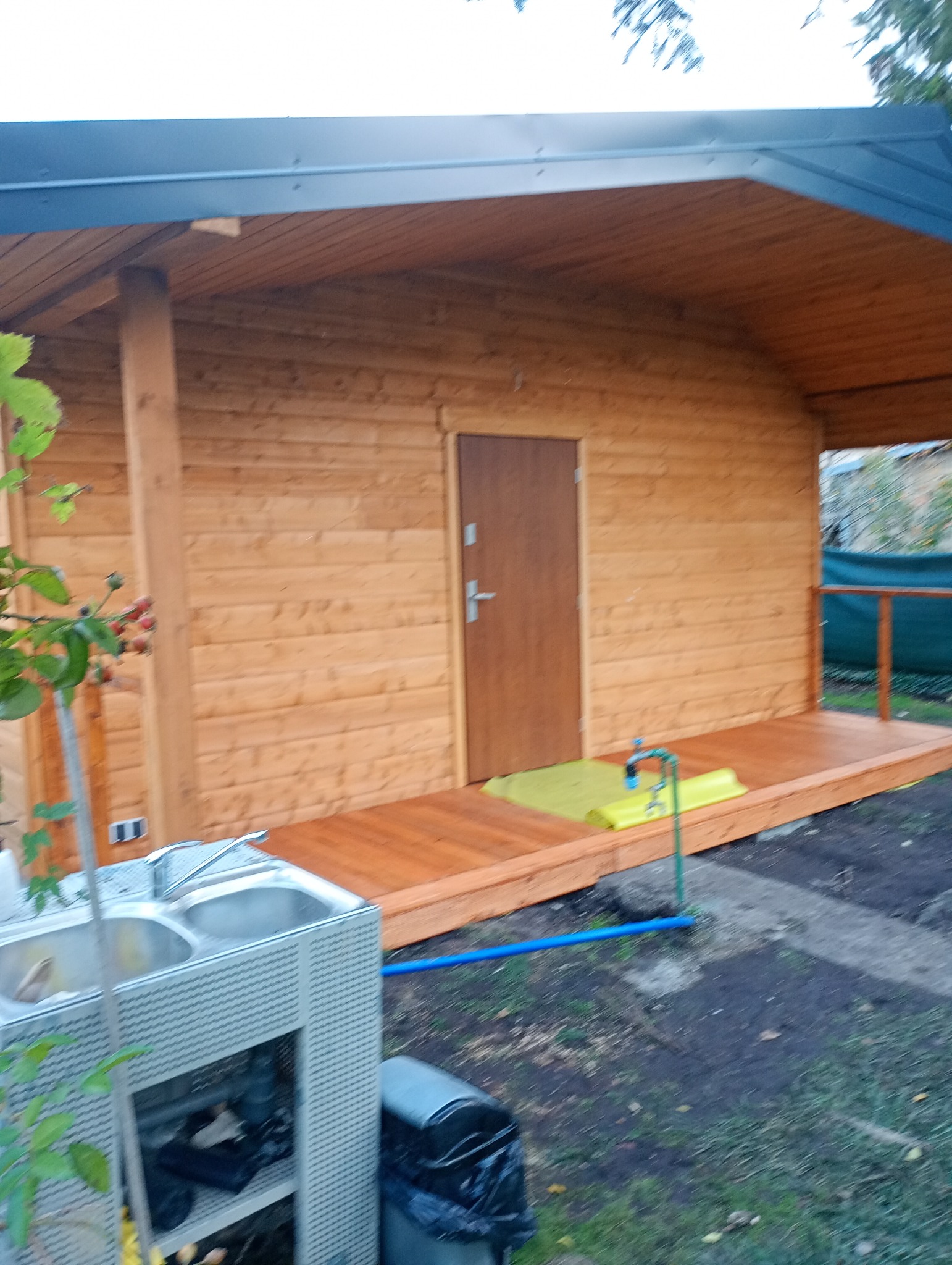 Drewniany domek letniskowy z tarasem, widok z zewnątrz. Widoczna instalacja wodna i zlewozmywak dwukomorowy. Realizacja w ogrodzie.