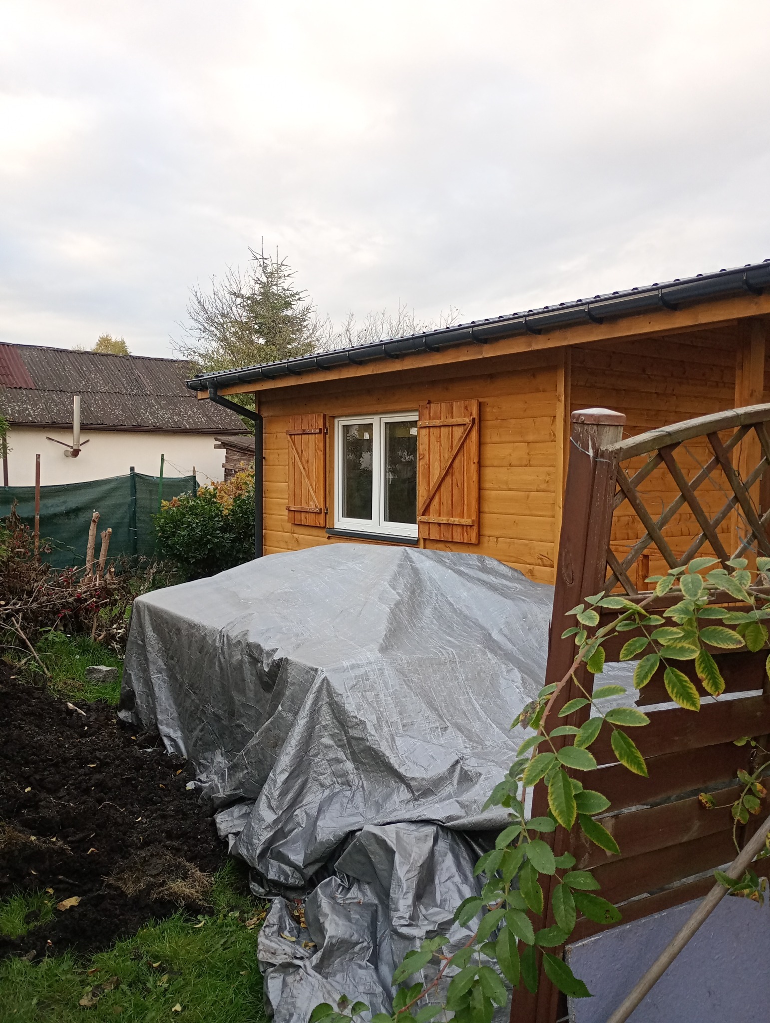 Drewniany domek z okiennicami, przykryty szarą plandeką na zewnątrz. Widoczne elementy ogrodu, drzewa i fragment dachu sąsiedniego budynku. Pochmurne niebo w tle.