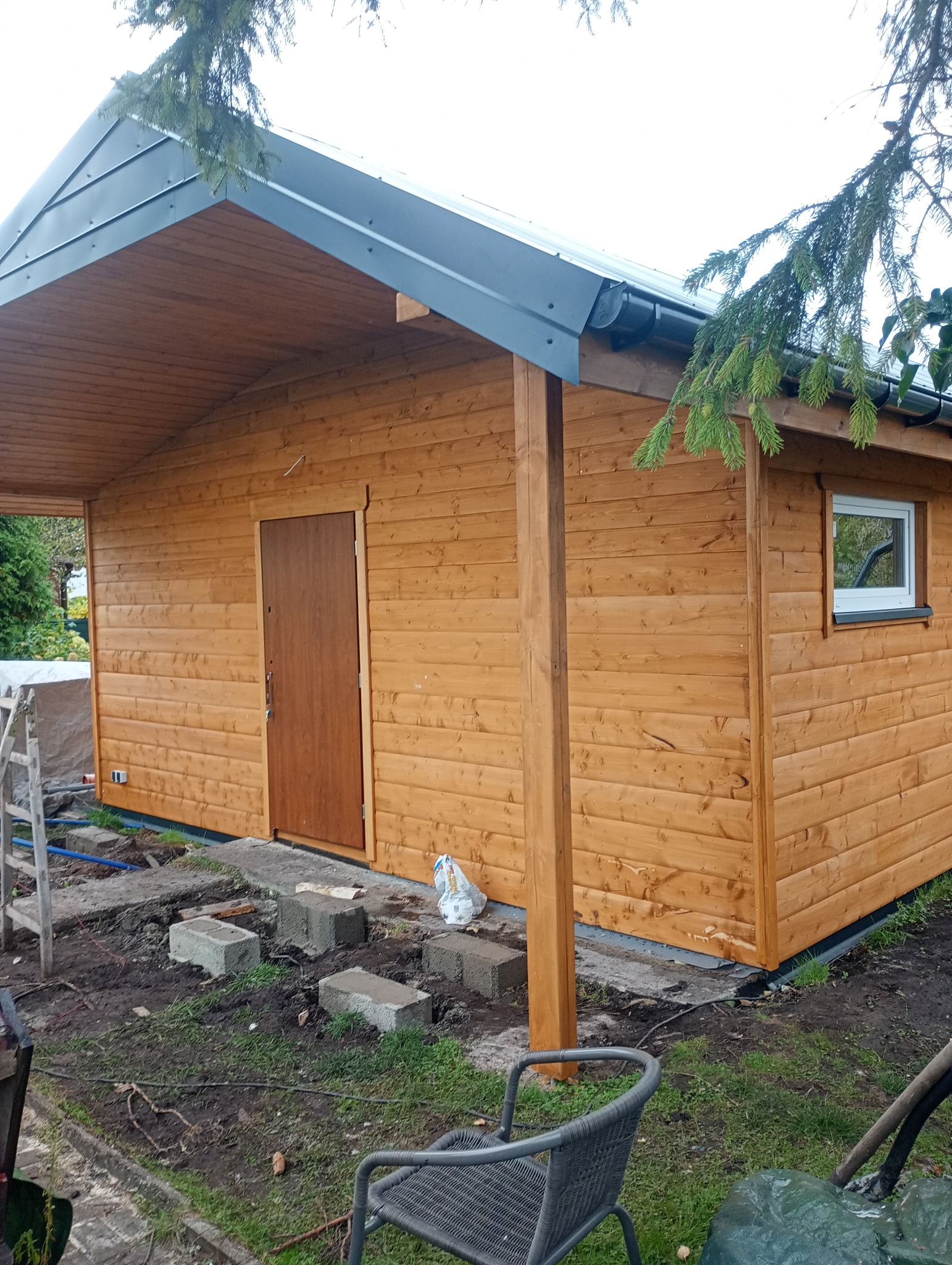 Drewniany domek z brązowymi drzwiami i szarym dachem, widoczny z zewnątrz, otoczony zielenią i elementami placu budowy. Świeżo pomalowany, w naturalnym odcieniu drewna.