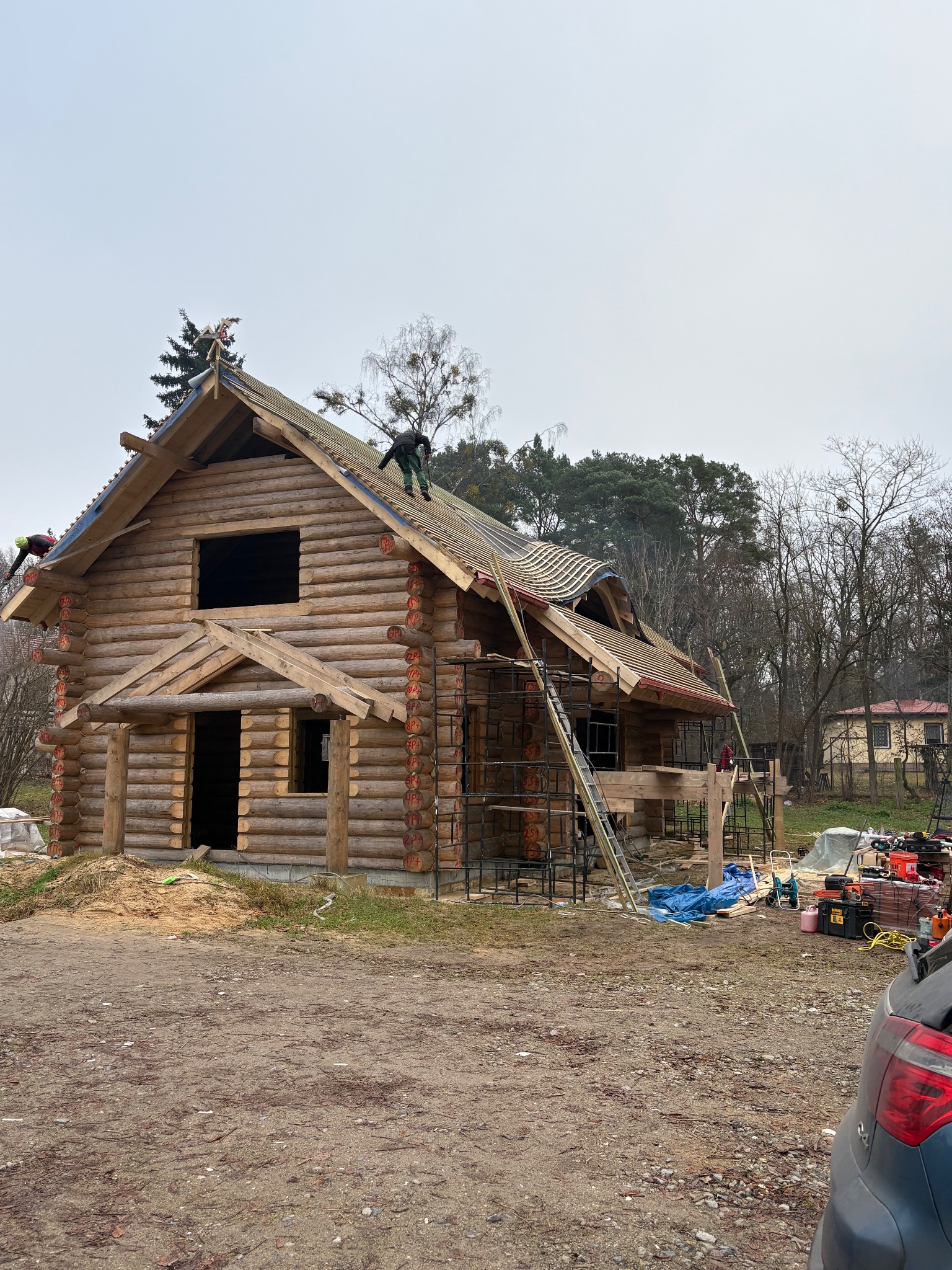 Budowa domu z bali w trakcie realizacji. Widoczni pracownicy na dachu, rusztowania i narzędzia. Drewniana konstrukcja z widocznymi oknami i drzwiami.