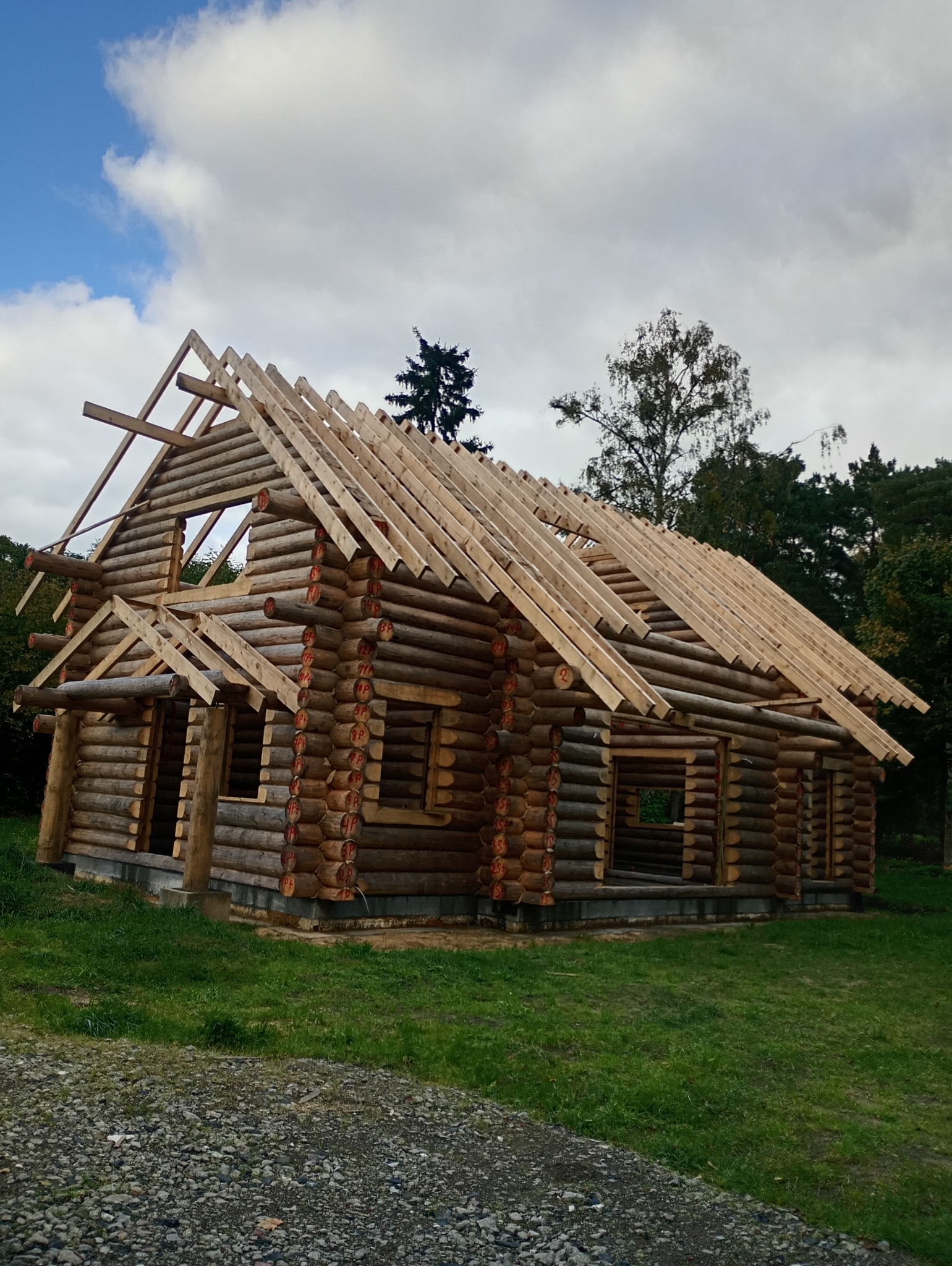 Surowy drewniany dom z bali w trakcie budowy, widoczne elementy konstrukcyjne dachu i numerowane bale, otoczony zielenią i częściowo wykończony fundament.