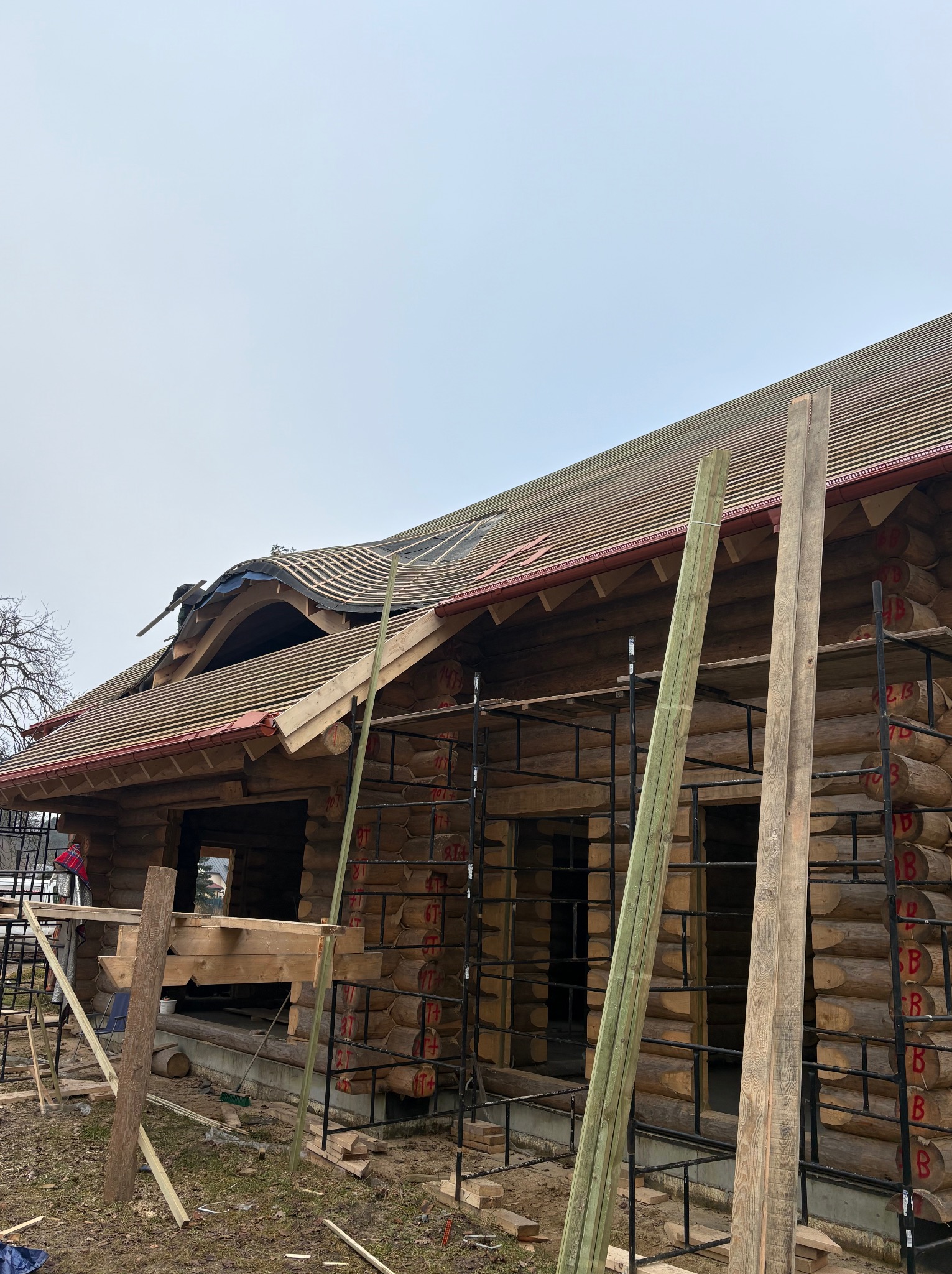 Budowa domu z bali: widok na konstrukcję dachu z deskami i rusztowaniami. Elementy drewniane z czerwonymi oznaczeniami. Prace w toku, pochmurne niebo w tle.