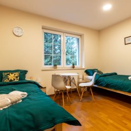 Ośrodek Terapii Uzależnień Poznaj Siebie - Pokój z dwoma pojedynczymi łóżkami z zieloną pościelą, stolikiem i krzesłami przy oknie, drewniana podłoga, zegar na ścianie.