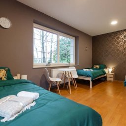 Ośrodek Terapii Uzależnień Poznaj Siebie - Pokój z trzema łóżkami pojedynczymi, nakrytymi zieloną pościelą i ręcznikami, w ośrodku w Koleczkowie. Na ścianie zegar i okno z widokiem na drzewa.