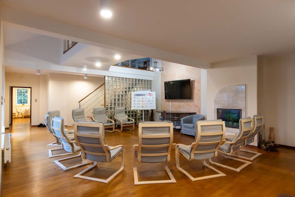 Jasne, przestronne wnętrze z krzesłami ustawionymi w okrąg, tablicą z napisem 'Program' i telewizorem na ścianie, prawdopodobnie sala do terapii grupowej.