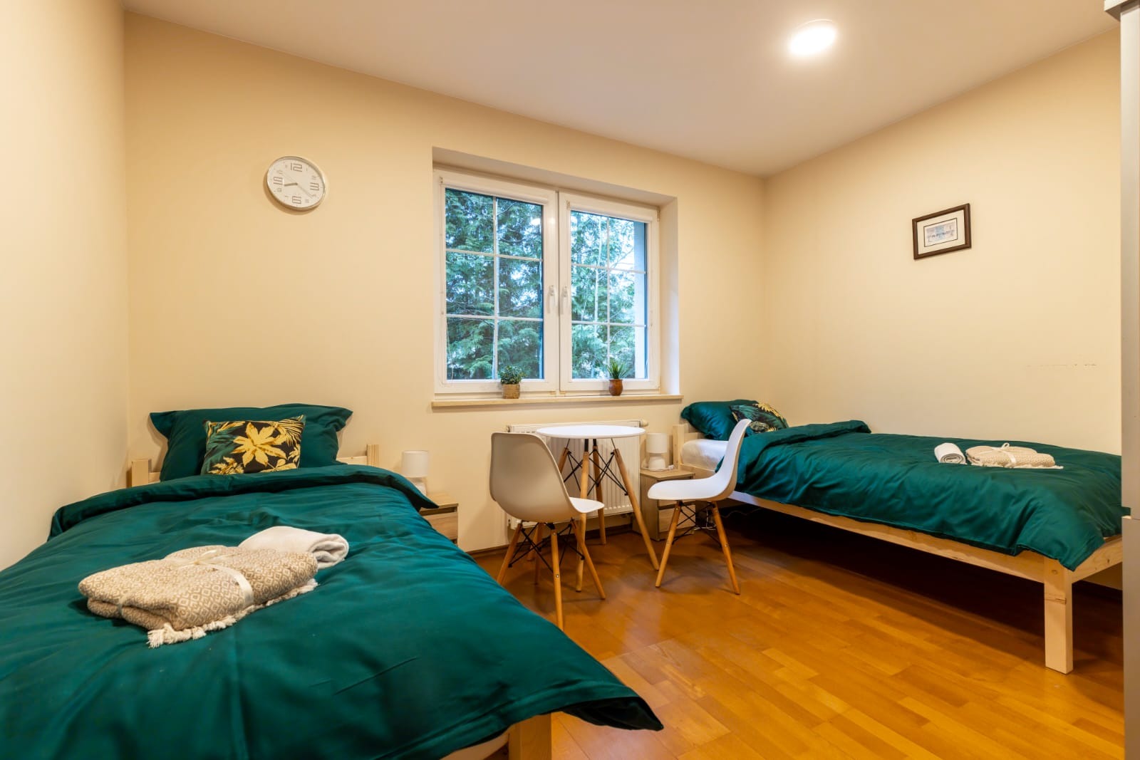 Pokój z dwoma pojedynczymi łóżkami z zieloną pościelą, stolikiem i krzesłami przy oknie, drewniana podłoga, zegar na ścianie.