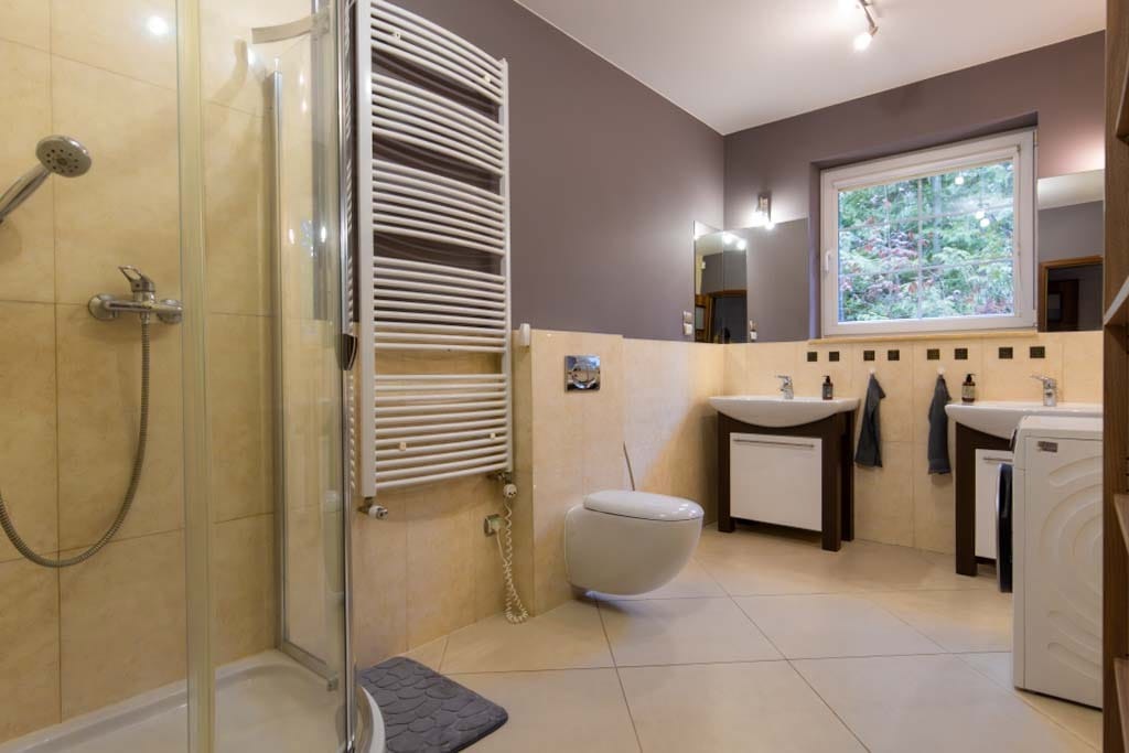 Nowoczesna łazienka z kabiną prysznicową, grzejnikiem drabinkowym, podwieszaną toaletą i dwiema umywalkami z szafkami w kolorach beżu i szarości.