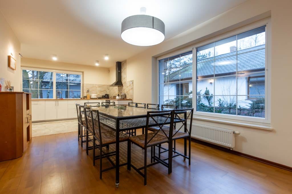 Jasne wnętrze domu z widokiem na ogród przez duże okno, stół z krzesłami w centralnej części, parkiet na podłodze, kuchnia w tle.