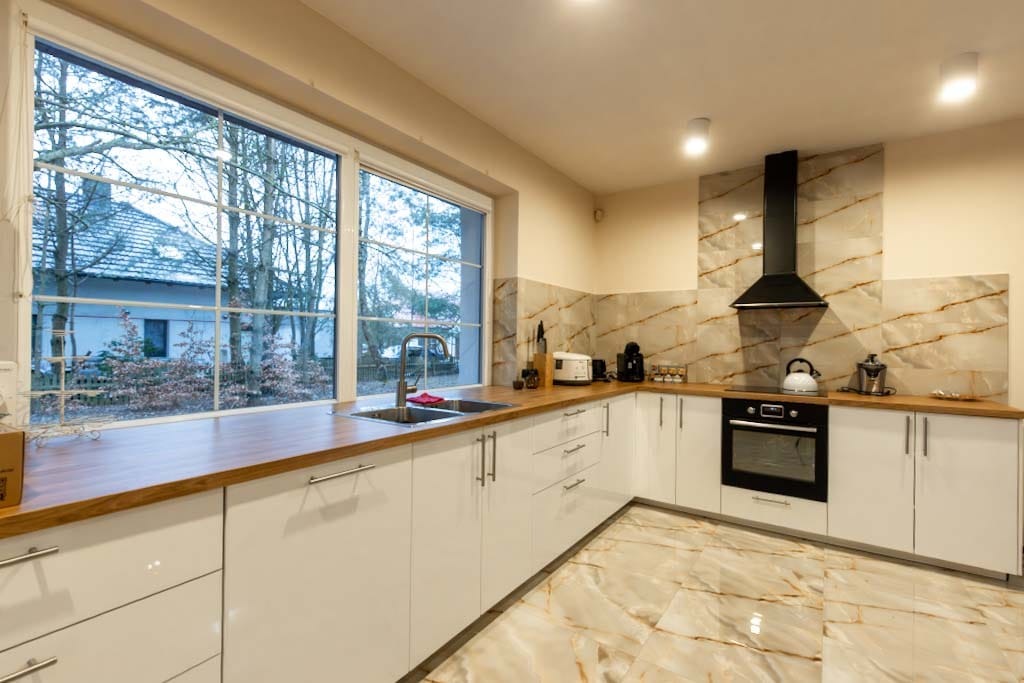 Nowoczesna, jasna kuchnia z białymi szafkami, drewnianym blatem i marmurową podłogą, widok na zimowy krajobraz za oknem.