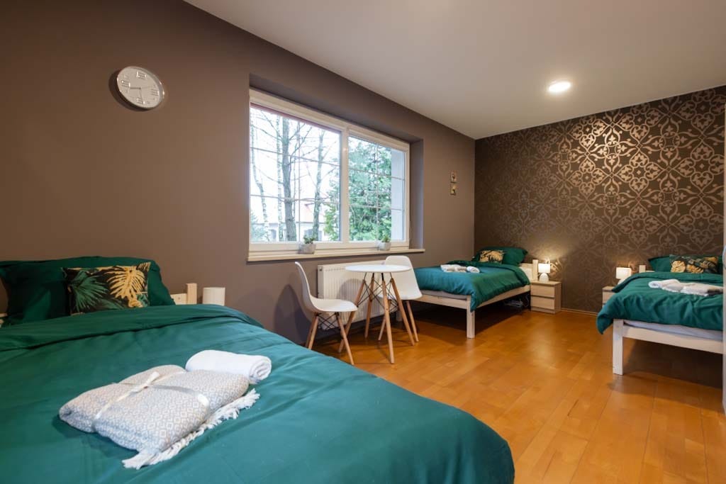 Pokój z trzema łóżkami pojedynczymi, nakrytymi zieloną pościelą i ręcznikami, w ośrodku w Koleczkowie. Na ścianie zegar i okno z widokiem na drzewa.