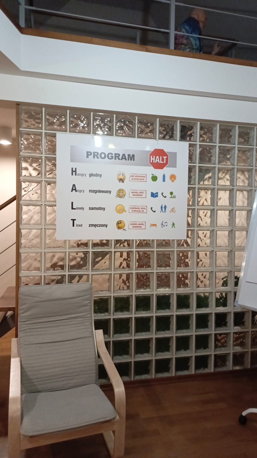 Plakat z programem HALT (głodny, rozgniewany, samotny, zmęczony) wiszący na ścianie z luksferów, szary fotel na pierwszym planie, osoba na balkonie w tle.