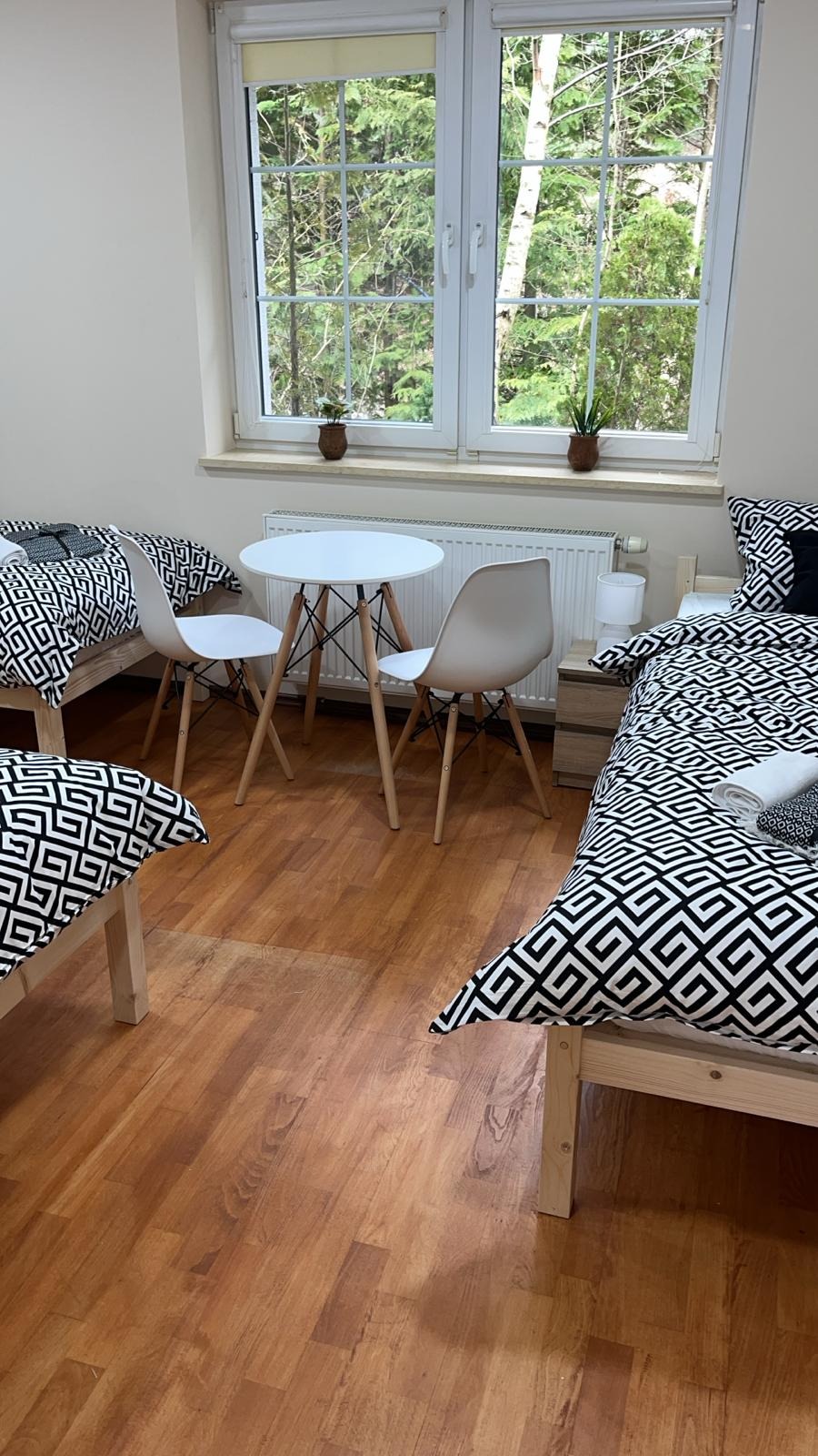 Wnętrze pokoju z trzema łóżkami w drewnianych ramach, pościel w geometryczne wzory, biały okrągły stolik z dwoma krzesłami, widok na las za oknem.