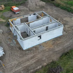 Surowy stan domu jednorodzinnego z pustaków, widok z drona, z widocznymi elementami konstrukcyjnymi, paletami i pomarańczową ciężarówką w tle.