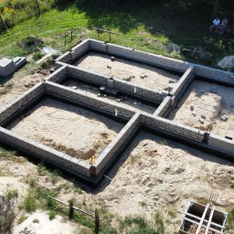 Paweł Czechowski - Widok z góry na fundamenty domu w trakcie budowy, widać bloczki betonowe tworzące ściany fundamentowe, piasek wewnątrz, teren budowy i osoby odpoczywające w tle.