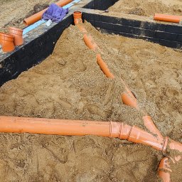 Paweł Czechowski - Układanie pomarańczowych rur kanalizacyjnych w wykopie fundamentowym, częściowo zasypanym piaskiem, z widocznymi izolowanymi ścianami fundamentów i pracownikiem łączącym elementy systemu.