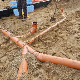 Paweł Czechowski - Pomarańczowe rury kanalizacyjne ułożone w piasku na budowie fundamentów, widoczne połączenia trójnikowe i kształtki, w tle pracownik w roboczych butach i fragment izolacji termicznej.