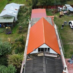 Paweł Czechowski - Widok z góry na dom w trakcie wymiany dachu na blachodachówkę w kolorze ceglastym, z widocznymi rusztowaniami, pracownikami i samochodem firmy budowlanej z reklamą na podwórku.