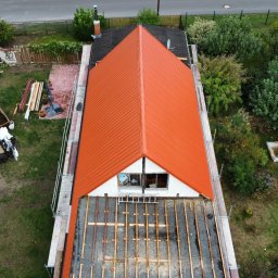 Paweł Czechowski - Widok z góry na dom w trakcie wymiany dachu z pomarańczową blachodachówką, widoczne rusztowania i elementy konstrukcji dachu, a w tle ogród z placem zabaw.