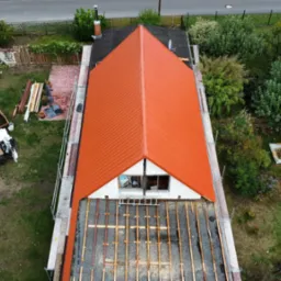 Widok z góry na dom w trakcie wymiany dachu z pomarańczową blachodachówką, widoczne rusztowania i elementy konstrukcji dachu, a w tle ogród z placem zabaw.