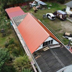 Paweł Czechowski - Widok z lotu ptaka na dom w trakcie wymiany dachu z czerwonej blachy, widoczne rusztowania, ekipa remontowa przy samochodzie firmowym i częściowo zdemontowany stary dach.
