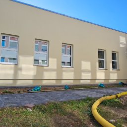 Paweł Czechowski - Elewacja budynku po malowaniu na jasny kremowy kolor, widoczne okna z białymi ramami, obok fragment budynku z drewnianą elewacją i żółtym obramowaniem wejścia, teren wokół budynku w trakcie prac...