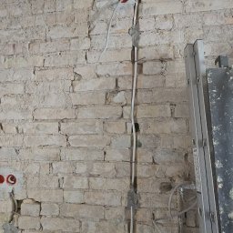 Paweł Czechowski - Surowa ściana z cegły z wystającymi przewodami elektrycznymi i puszkami, częściowo zamaskowanymi szarym cementem, obok stoi drabina.
