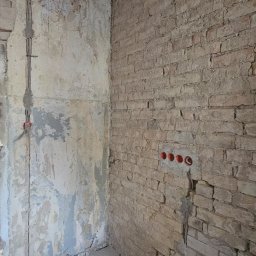 Paweł Czechowski - Wnętrze pokoju w trakcie remontu: surowa ceglana ściana z instalacją elektryczną, otynkowana ściana z widocznymi śladami farby, odsłonięty drewniany strop i betonowa podłoga.