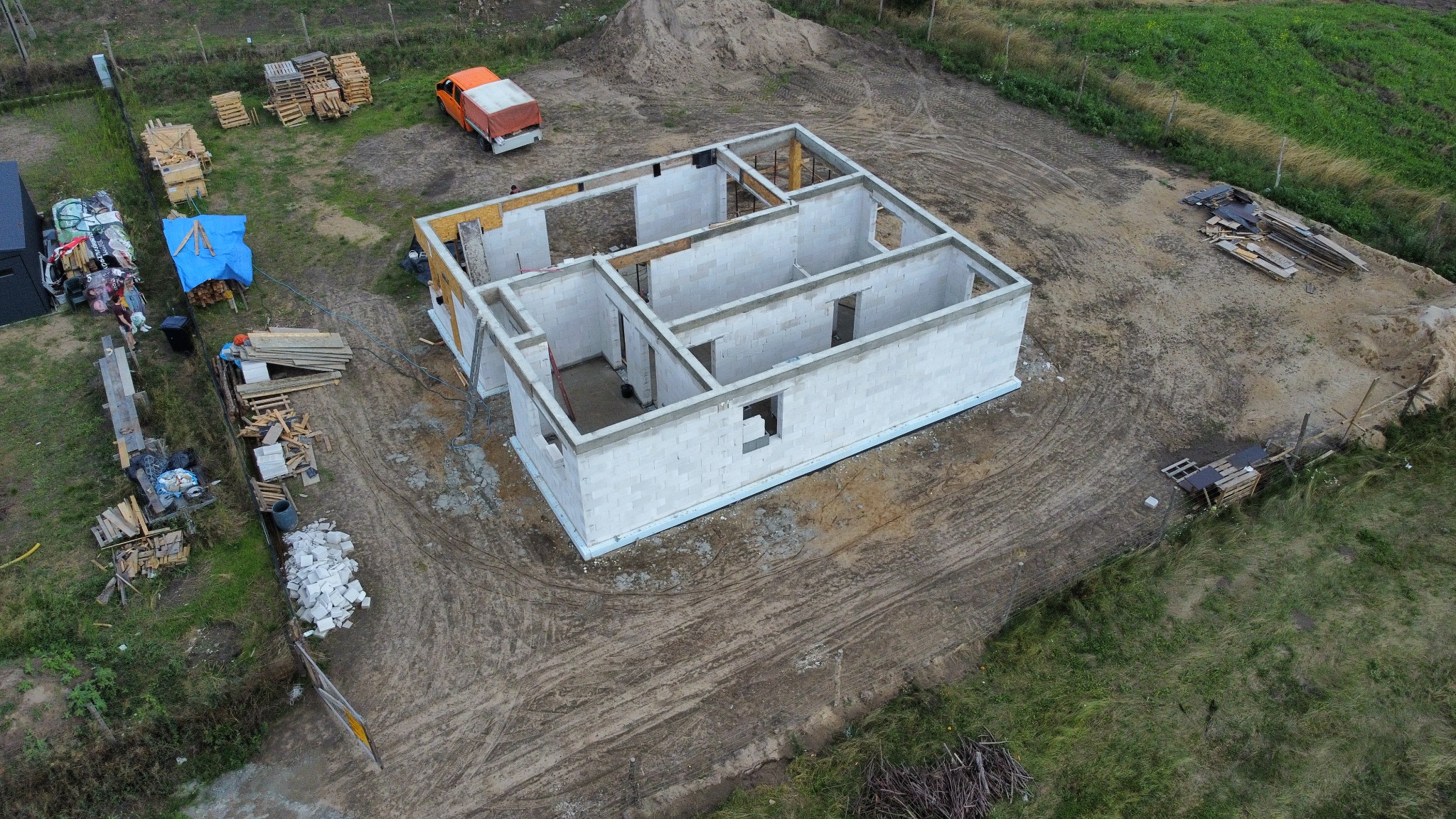 Surowy stan domu jednorodzinnego z pustaków, widok z drona, z widocznymi elementami konstrukcyjnymi, paletami i pomarańczową ciężarówką w tle.