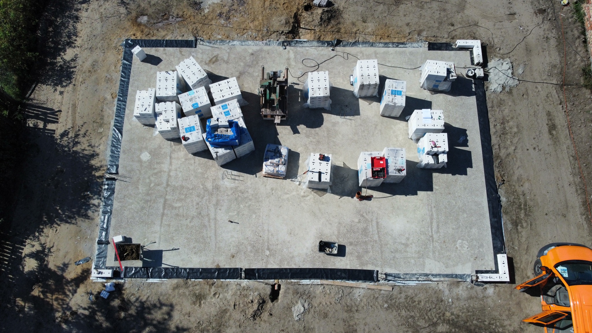 Widok z góry na przygotowany teren pod fundamenty, z wyznaczonym obrysem i rozłożonymi bloczkami betonowymi, obok stoi pomarańczowy samochód dostawczy z otwartymi drzwiami.
